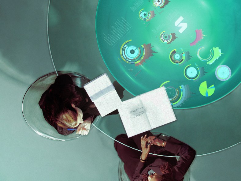 Two businesswomen in discussion at glass table, overhead view